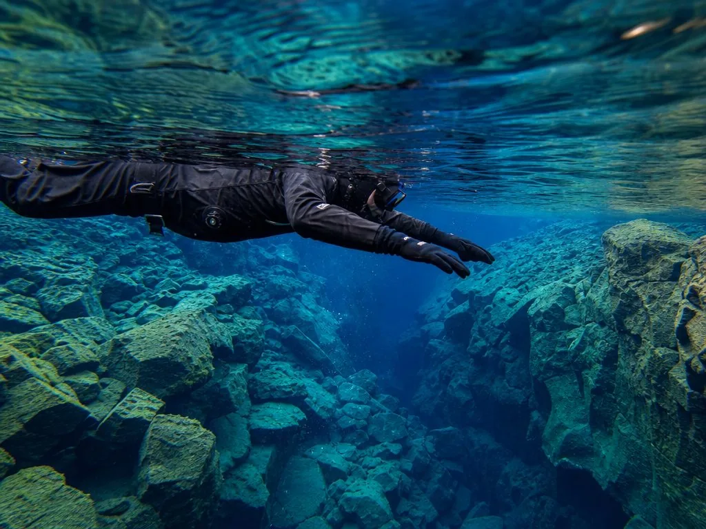 A snorkeler in a drysuit and hood, hood and gloves visible, floatng over the rift